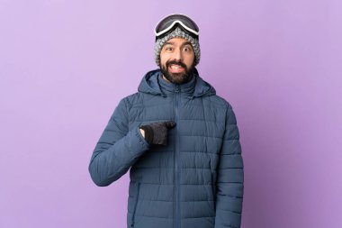 İzole edilmiş mor arka planda snowboard gözlüklü kayakçı kendini işaret ediyor.