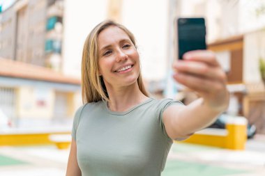 Açık havada genç sarışın bir kadın cep telefonuyla selfie çekiyor.