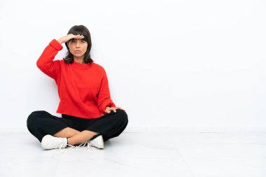 Genç, melez bir kadın, beyaz arka planda izole bir şekilde yerde oturuyor. Bir şeye bakmak için uzaklara bakıyor.