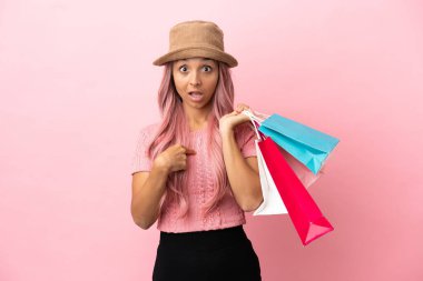 Alışveriş çantalı genç melez kadın pembe arka planda izole edilmiş, şaşırmış bir yüz ifadesi var.
