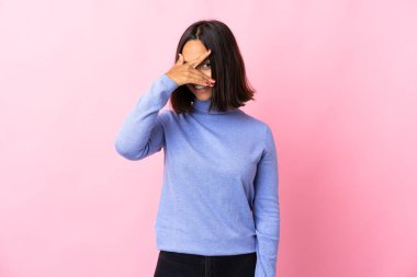 Pembe arka planda izole edilmiş genç Latin bir kadın. Gözleri elleriyle kaplıyor ve gülümsüyor.