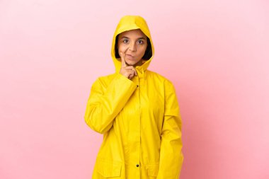 Yağmura dayanıklı bir ceket giyen genç Latin bir kadın izole edilmiş arka planda düşünüyor.