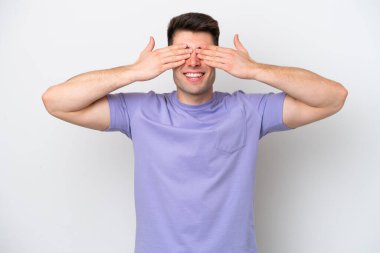 Beyaz arka planda izole edilmiş beyaz tenli genç bir adam. Gözleri elleriyle kaplıyor ve gülümsüyor.