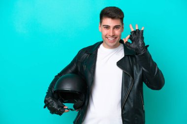 Mavi arka planda izole edilmiş motosiklet kasklı genç beyaz adam parmaklarıyla işaret veriyor.