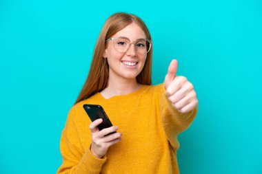 Genç kızıl saçlı kadın mavi arka planda izole edilmiş. Baş parmak kullanırken cep telefonu kullanıyor.