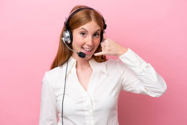 Telemarketer redhead woman working with a headset isolated on pink background making phone gesture. Call me back sign clipart