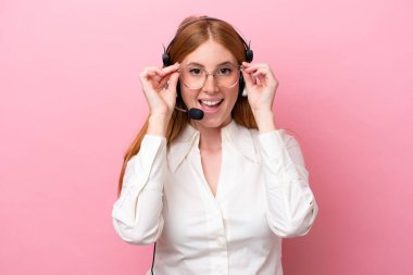 Pembe arka planda gözlüklü ve şaşırmış bir kulaklıkla çalışan pazarlamacı kızıl saçlı kadın.