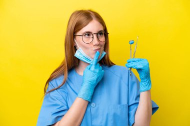 Genç kızıl saçlı dişçi kadın sarı arka planda izole edilmiş, araştırırken şüpheleri var.