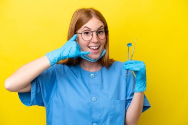Genç kızıl saçlı dişçi kadın sarı arka planda izole edilmiş telefon jesti yapıyor. Bana geri dönüş işareti ver.