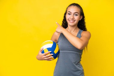 Young woman playing volleyball isolated on yellow background pointing back