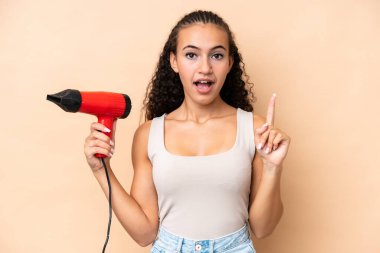 Elinde bej arka planda izole edilmiş saç kurutma makinesi tutan genç bir kadın çözümü bulmak için parmağını kaldırıyor.