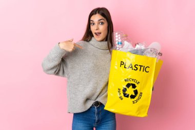 Elinde plastik şişelerle dolu bir çanta tutan beyaz bir kadın. Pembe arka planda izole edilmiş, şaşırmış bir yüz ifadesiyle geri dönüşüm için.