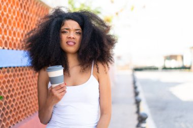 Genç Afrikalı Amerikalı bir kadın, elinde üzgün bir ifadeyle açık havada kahve tutuyor.