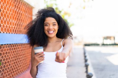 Genç Afrikalı Amerikalı bir kadın açık havada kahvesini yudumlarken kendine güvenen bir ifadeyle seni işaret ediyor.