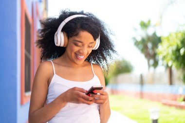 Dışarıda kulaklıklı genç Afrikalı Amerikalı kadın cep telefonuyla mesaj yolluyor.