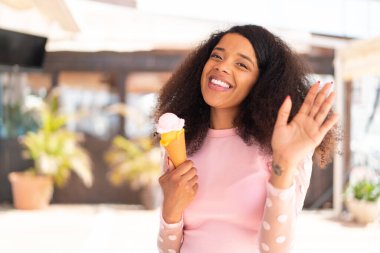 Dışarıda elinde kornet dondurmasıyla genç Afrikalı Amerikalı kadın mutlu bir ifadeyle selamlıyor.