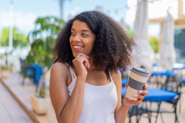 Genç Afrikalı Amerikalı bir kadın açık havada kahvesini yudumlarken bir fikir üretiyor ve etrafına bakıyor.