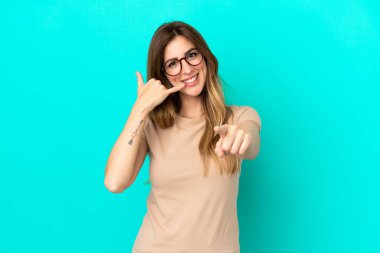 Mavi arka planda izole edilmiş genç beyaz kadın telefon hareketlerinde bulunuyor ve ön tarafı işaret ediyor.