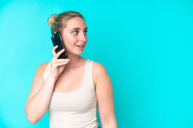 Mavi arka planda izole edilmiş genç beyaz bir kadın cep telefonuyla sohbet ediyor.