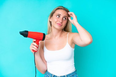 Young caucasian woman holding a hairdryer isolated on blue background having doubts and with confuse face expression