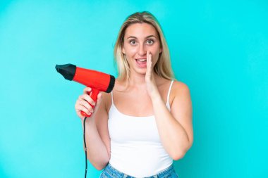 Elinde saç kurutma makinesi olan beyaz bir kadın mavi arka planda ağzı açık bağırıyor.