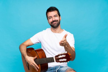 İzole edilmiş mavi arka planda gitar çalan genç adam iyi bir anlaşma için el sıkışıyor.