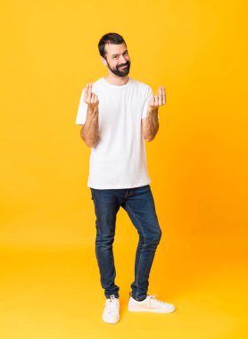 Sakallı, izole edilmiş sarı arka planda para hareketi yapan bir adamın tam boy fotoğrafı.