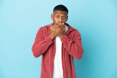 Mavi arka planda izole edilmiş genç latin adam elleriyle ağzını kapatıyor.