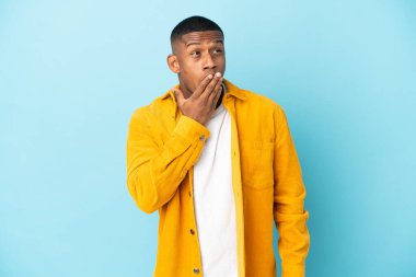 Genç latin adam mavi arka planda izole edilmiş esniyor ve ağzını eliyle açık tutuyor.