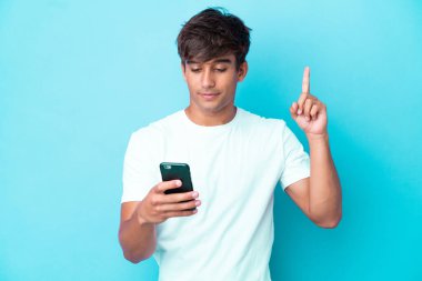Genç beyaz adam mavi arka planda cep telefonu ve parmak kaldırarak izole edildi.