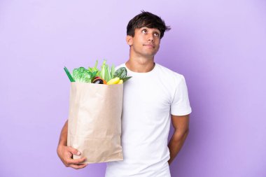 Young man holding a grocery shopping bag isolated on purple background and looking up