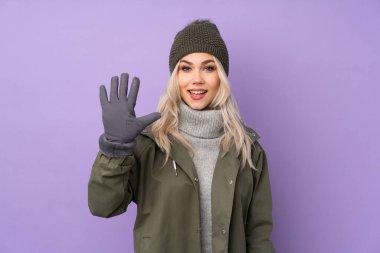 Kışlık şapkalı genç sarışın kız izole edilmiş mor arka planda parmaklarıyla beşi sayıyor.