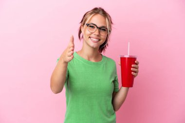 Pembe arka planda izole edilmiş bir içecek tutan genç Rus kadın iyi bir anlaşma için el sıkışıyor.