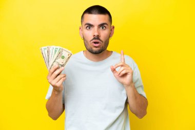 Genç beyaz adam sarı arka planda izole edilmiş bir sürü para alıyor. Çözümü bulmak için parmağını kaldırıyor.