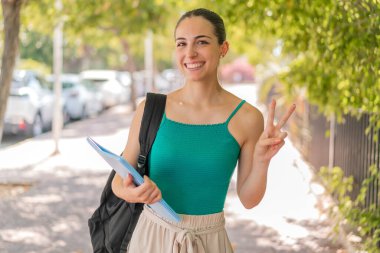 Açık havada gülümseyen ve zafer işareti gösteren genç ve güzel bir bayan öğrenci.