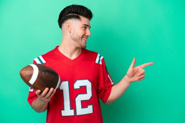 Rugby oynayan beyaz bir genç, yeşil arka planda izole edilmiş bir şekilde parmak göstererek bir ürün sunuyor.