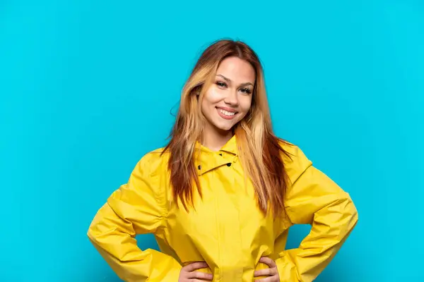 stock image Teenager girl wearing a rainproof coat over isolated blue background posing with arms at hip and smiling