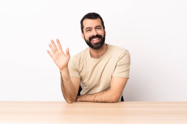 Masada sakallı, beyaz bir adam elinde mutlu bir ifadeyle selam veriyor..