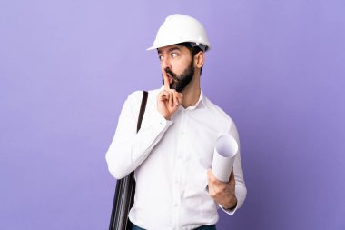 Miğferli genç bir mimar, izole edilmiş mor arkaplanının planlarını tutuyor. Parmağını ağzına koyduğunda sessizlik işareti gösteriyor.