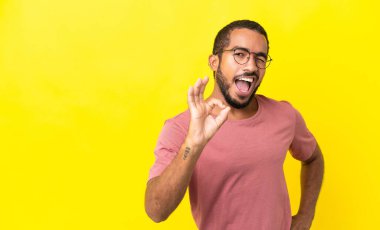 Sarı arka planda izole edilmiş genç Latin adam parmaklarıyla tamam işareti gösteriyor.