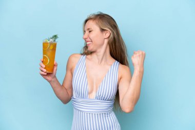 Mavi arka planda bir kokteyl tutan mayo giyen genç sarışın bir kadın zaferi kutluyor.