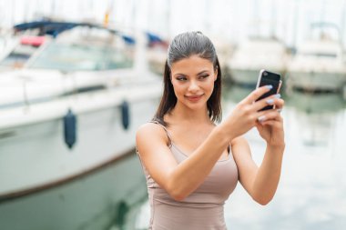 Açık havada genç ve güzel bir kadın cep telefonuyla selfie çekiyor.