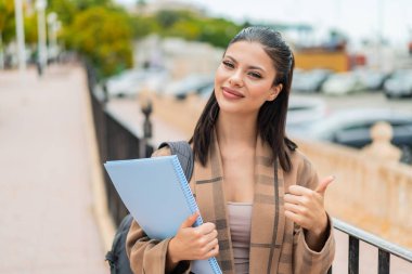 Açık havada genç ve güzel bir kız öğrenci. Başparmakları havada. Çünkü iyi bir şey oldu.