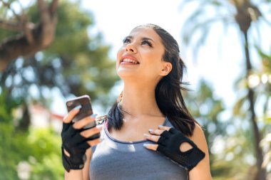 Dışarıda cep telefonu kullanan genç ve güzel bir bayan gülümserken yukarı bakıyor.