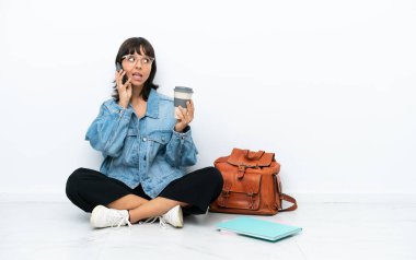 Genç bir kız öğrenci beyaz arka planda tek başına oturuyor. Elinde kahve ve cep telefonu var.