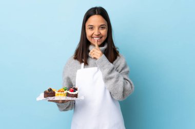 Pasta şefi, izole edilmiş mavi arka planda büyük bir pasta tutuyor. Parmağını ağzına koyduğunda sessizlik işareti gösteriyor..