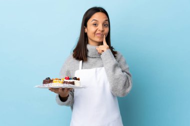 Pasta şefi elinde büyük bir pasta tutarak izole edilmiş mavi arka planda düşünüyor..