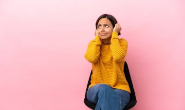 Genç melez bir kadın pembe arka planda izole edilmiş bir sandalyede oturuyor hayal kırıklığına uğramış ve kulaklarını kapatmış.