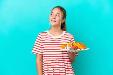 Mavi arka planda gözleme tutan küçük beyaz kız yukarı bakarken bir fikir düşünüyor.