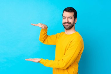 İzole edilmiş mavi arka plan üzerinde genç beyaz bir adam ilan vermek için kopyalama alanı tutuyor.